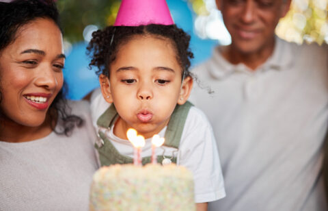 Child wearing a party hat blowing out birthday candles on a cake while parents smile nearby