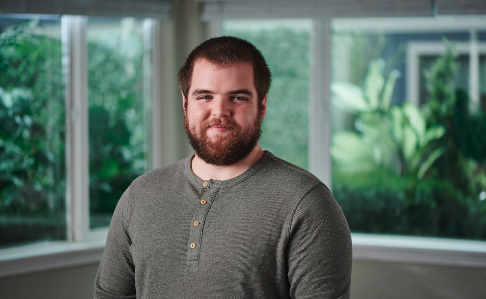 Dalton smiles at the camera while standing indoors near large windows with greenery outside.