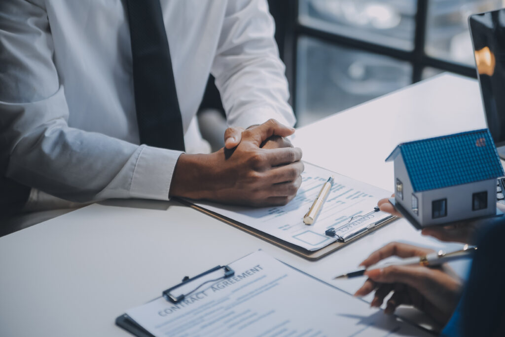 Close-up of two people at a desk with contract paperwork, a model house, and a pen