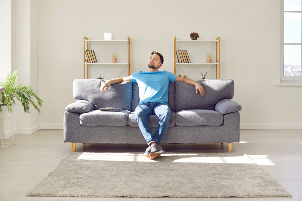 Relaxed young man is enjoying free time and sitting on comfortable couch at home