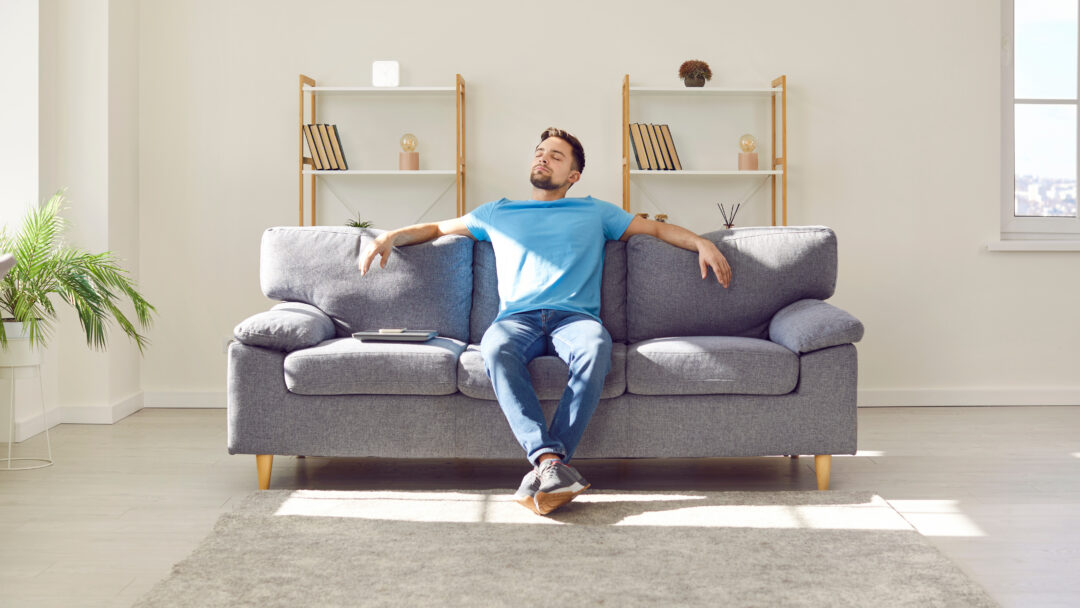 Relaxed young man is enjoying free time and sitting on comfortable couch at home