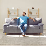 Relaxed young man is enjoying free time and sitting on comfortable couch at home