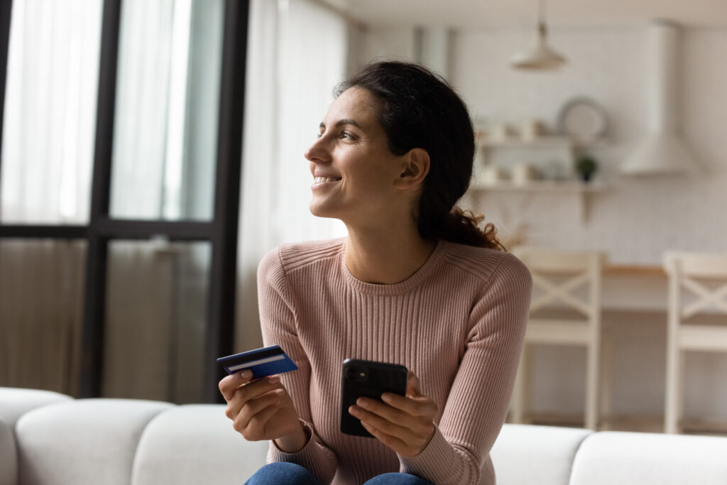Woman holding a credit card and smartphone while sitting on a couch, smiling as she shops or manages finances online.
