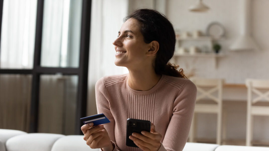 Woman holding a credit card and smartphone while sitting on a couch, smiling as she shops or manages finances online.