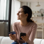 Woman holding a credit card and smartphone while sitting on a couch, smiling as she shops or manages finances online.