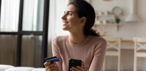 Woman holding a credit card and smartphone while sitting on a couch, smiling as she shops or manages finances online.