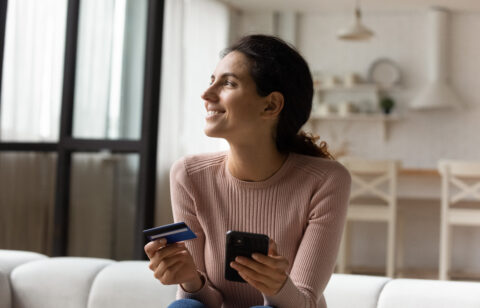 Woman holding a credit card and smartphone while sitting on a couch, smiling as she shops or manages finances online.