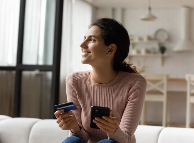 Woman holding a credit card and smartphone while sitting on a couch, smiling as she shops or manages finances online.