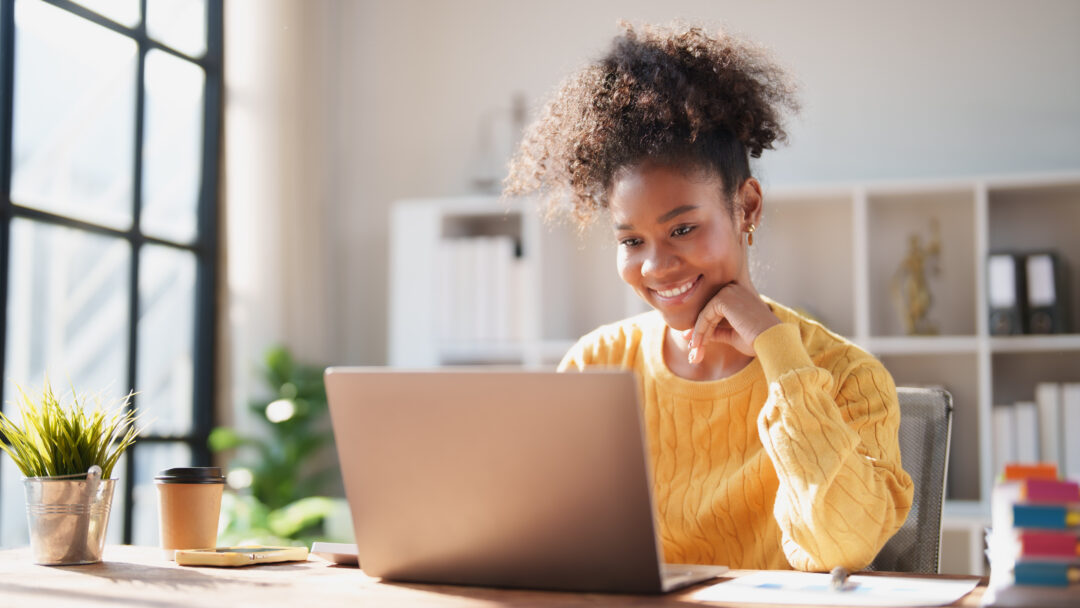 Young happy African American woman smiling while looking at a laptop screen, engaging in online work from a modern office, enjoying remote productivity