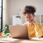 Young happy African American woman smiling while looking at a laptop screen, engaging in online work from a modern office, enjoying remote productivity