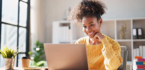 Young happy African American woman smiling while looking at a laptop screen, engaging in online work from a modern office, enjoying remote productivity
