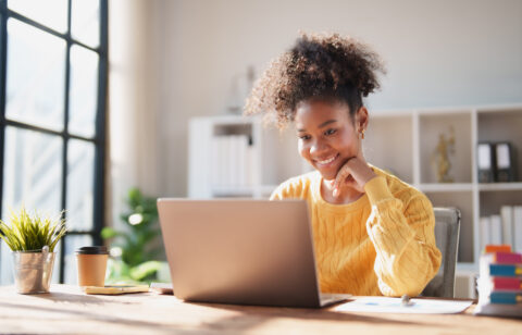 Young happy African American woman smiling while looking at a laptop screen, engaging in online work from a modern office, enjoying remote productivity