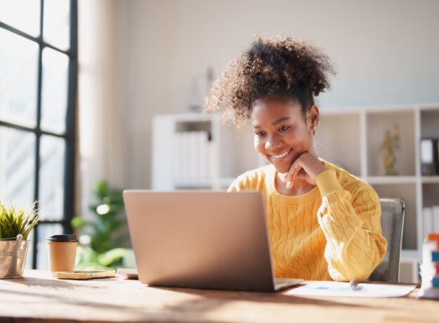 Young happy African American woman smiling while looking at a laptop screen, engaging in online work from a modern office, enjoying remote productivity