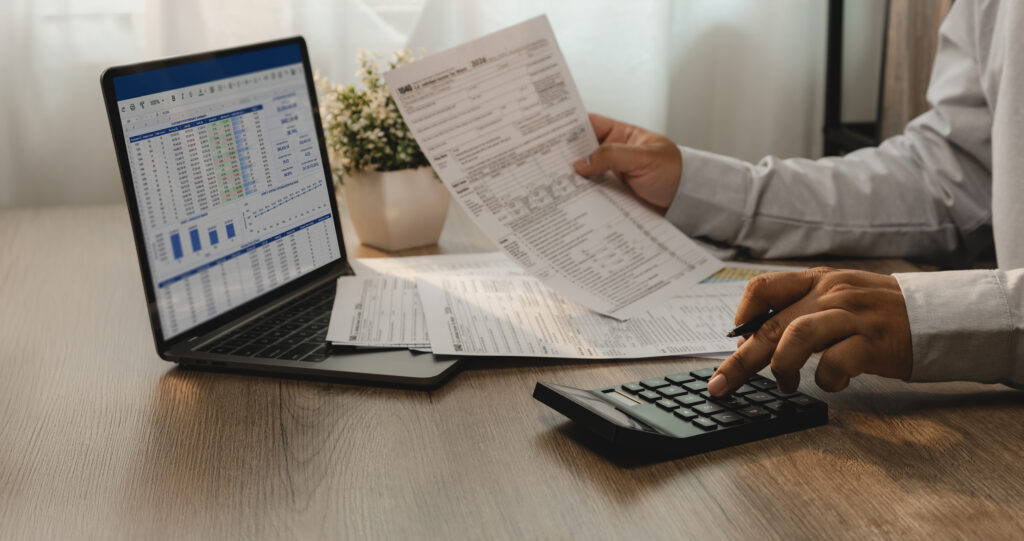 Person reviewing financial documents while using a calculator and laptop displaying a spreadsheet.