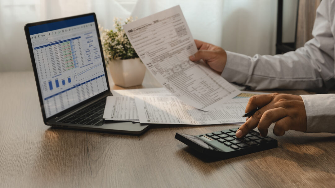 Person reviewing financial documents while using a calculator and laptop displaying a spreadsheet.