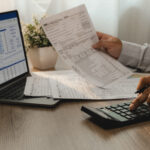 Person reviewing financial documents while using a calculator and laptop displaying a spreadsheet.