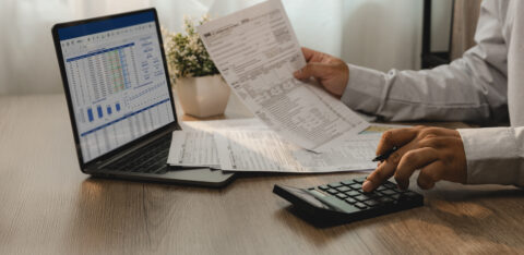Person reviewing financial documents while using a calculator and laptop displaying a spreadsheet.