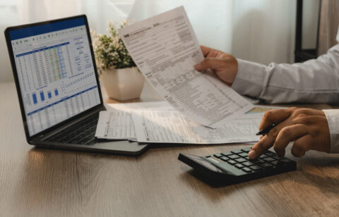 Person reviewing financial documents while using a calculator and laptop displaying a spreadsheet.