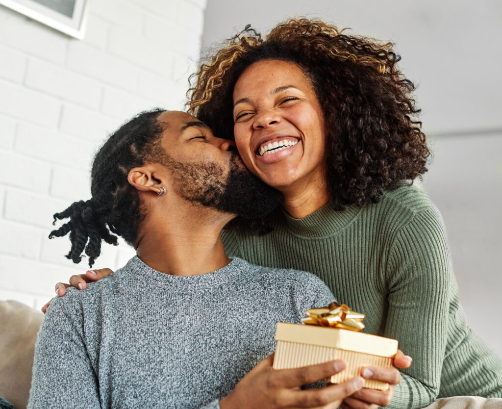 Man holding a wrapped gift while kissing a smiling woman on the cheek in a cozy home setting.