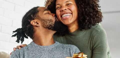 Man holding a wrapped gift while kissing a smiling woman on the cheek in a cozy home setting.