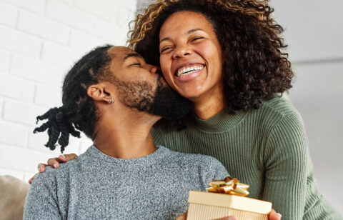Man holding a wrapped gift while kissing a smiling woman on the cheek in a cozy home setting.