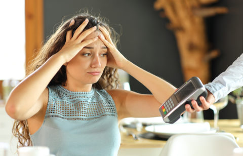 A worried person throws their hands on her head as a restaurant server extends a payment terminal toward them to pay the bill.