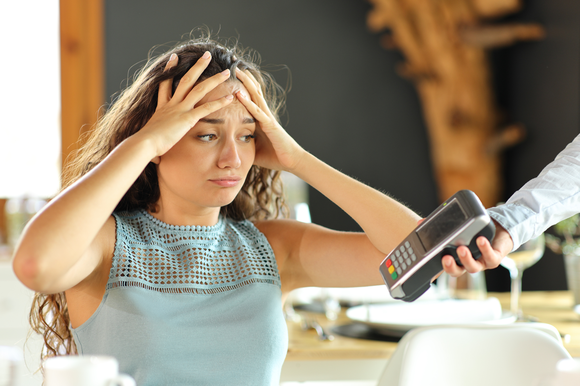 A worried person throws their hands on her head as a restaurant server extends a payment terminal toward them to pay the bill.