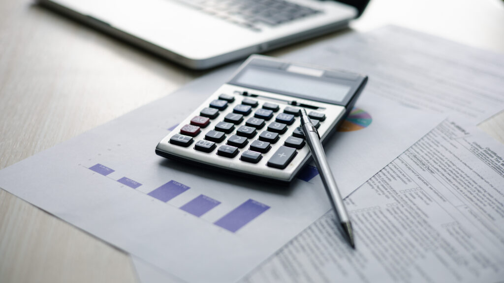 Calculator and pen resting on financial reports and charts beside a laptop.