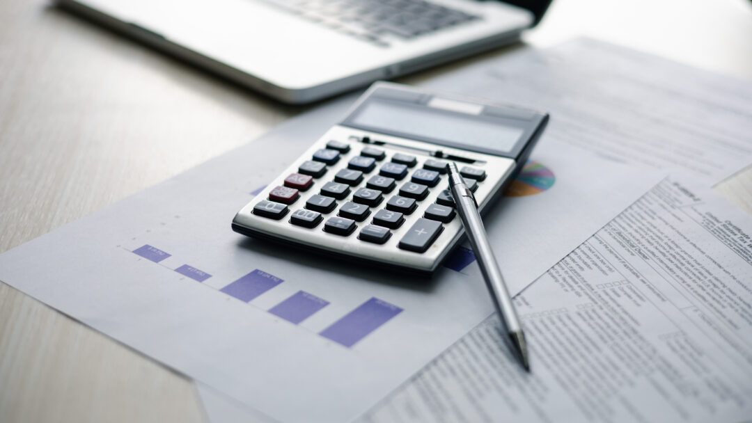 Calculator and pen resting on financial reports and charts beside a laptop.