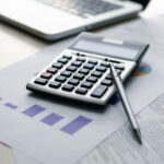 Calculator and pen resting on financial reports and charts beside a laptop.