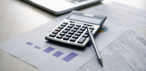 Calculator and pen resting on financial reports and charts beside a laptop.