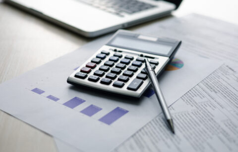 Calculator and pen resting on financial reports and charts beside a laptop.
