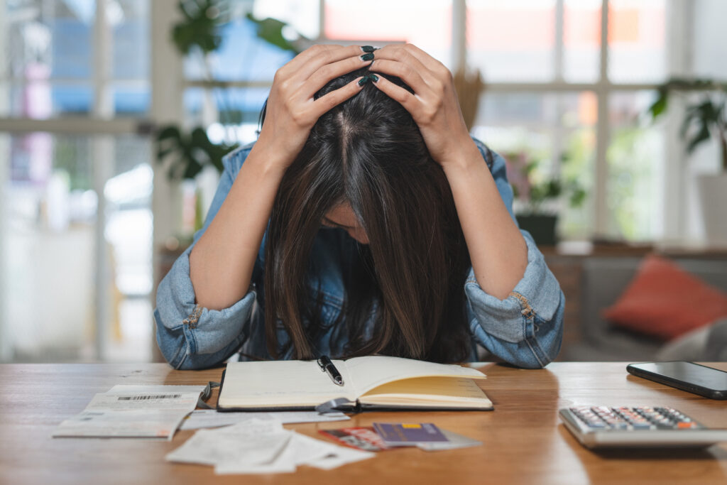 Stressed woman holding her head while sitting at a table with bills, receipts, credit cards, a calculator, and a notebook.