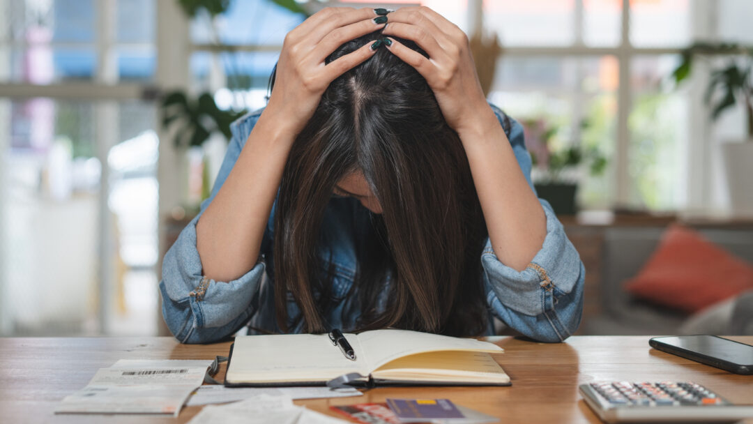 Stressed woman holding her head while sitting at a table with bills, receipts, credit cards, a calculator, and a notebook.