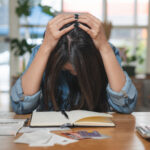 Stressed woman holding her head while sitting at a table with bills, receipts, credit cards, a calculator, and a notebook.