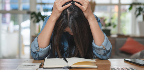 Stressed woman holding her head while sitting at a table with bills, receipts, credit cards, a calculator, and a notebook.