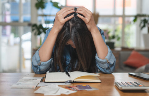 Stressed woman holding her head while sitting at a table with bills, receipts, credit cards, a calculator, and a notebook.