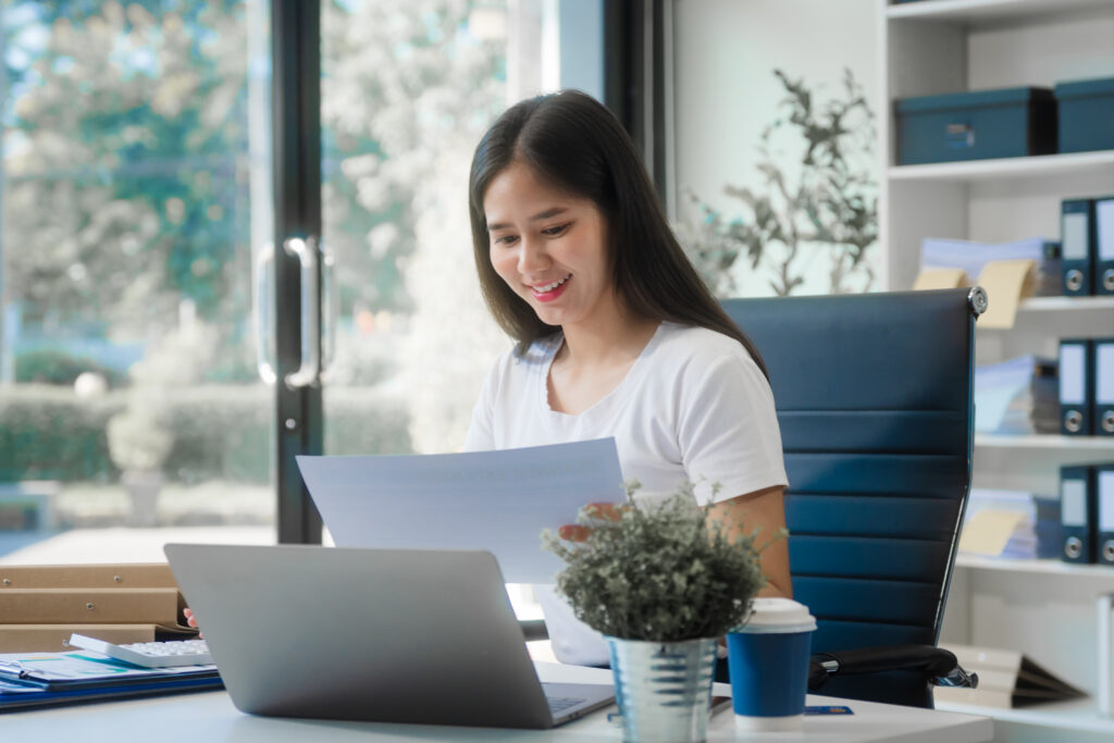 Woman reviewing financial documents at her desk with a laptop in a bright office, smiling while checking paperwork.