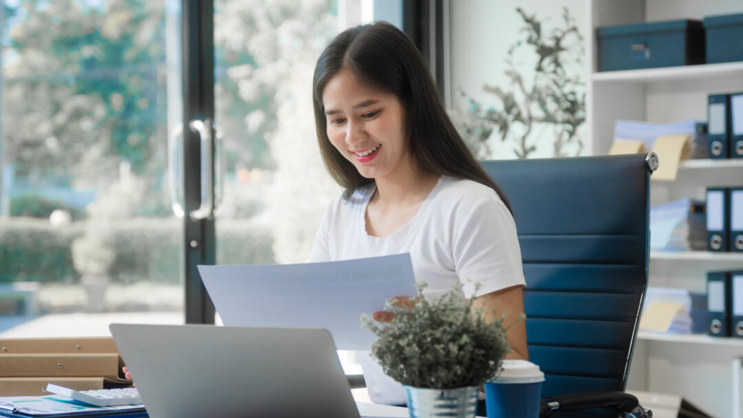 Woman reviewing financial documents at her desk with a laptop in a bright office, smiling while checking paperwork.