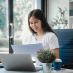 Woman reviewing financial documents at her desk with a laptop in a bright office, smiling while checking paperwork.