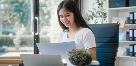 Woman reviewing financial documents at her desk with a laptop in a bright office, smiling while checking paperwork.