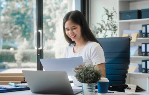 Woman reviewing financial documents at her desk with a laptop in a bright office, smiling while checking paperwork.