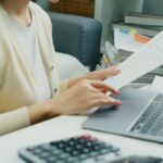 Individual Reviewing Financial Reports.An Asian individual reviews financial reports and property rates, calculating income and expenses with serious dedication using technology.