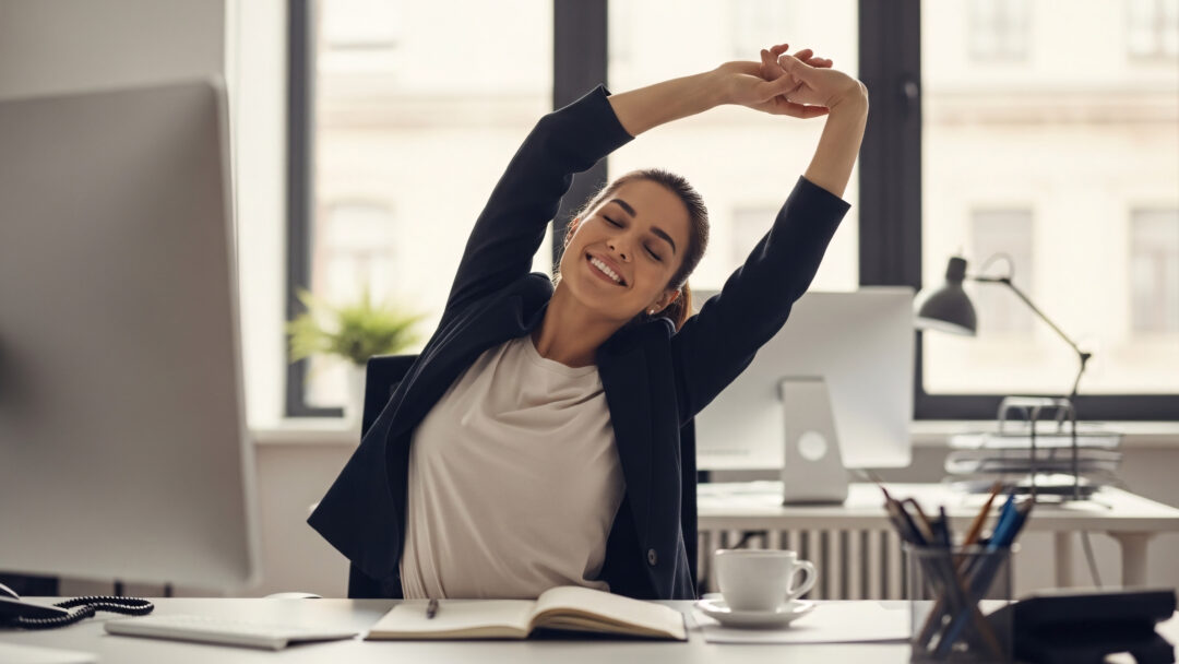 Woman stretching at her desk in an office while taking a break from working on her computer.