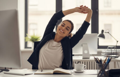 Woman stretching at her desk in an office while taking a break from working on her computer.
