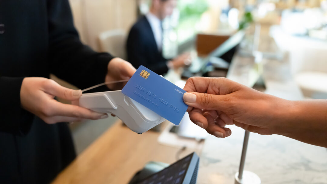 A customer holds a blue credit card near a contactless payment terminal at a checkout counter.