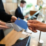 A customer holds a blue credit card near a contactless payment terminal at a checkout counter.