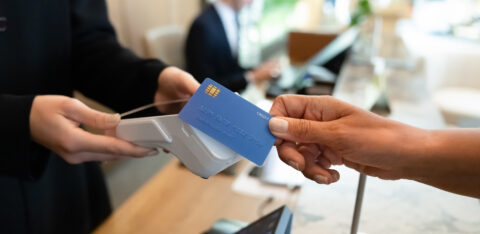 A customer holds a blue credit card near a contactless payment terminal at a checkout counter.