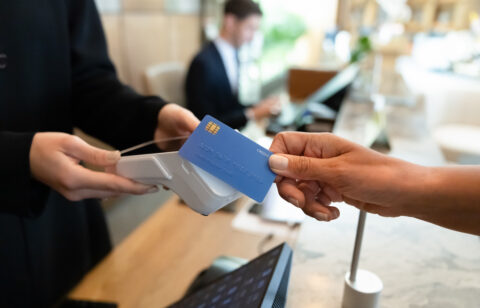 A customer holds a blue credit card near a contactless payment terminal at a checkout counter.