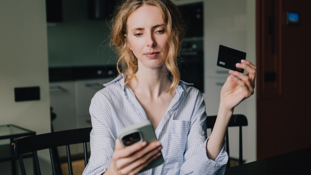 A woman holds a credit card while looking at her smartphone at home.